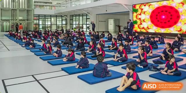 A large group of Merchiston International School Shenzhen students sitting on blue exercise mats in a gymnasium for a fitness or assembly session.