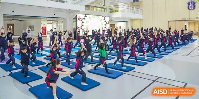 A very large group of Merchiston International School Shenzhen students actively participating in a group fitness or exercise routine in the school hall.