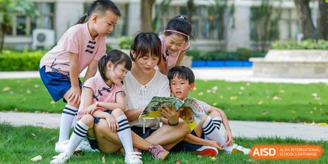 A teacher from Guangdong Country Garden School reads a book to two young children on a green lawn.