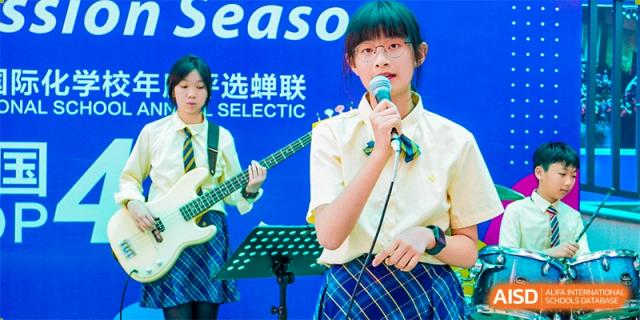 A female student from Guangdong Country Garden School in a plaid uniform sings into a microphone during a band performance.