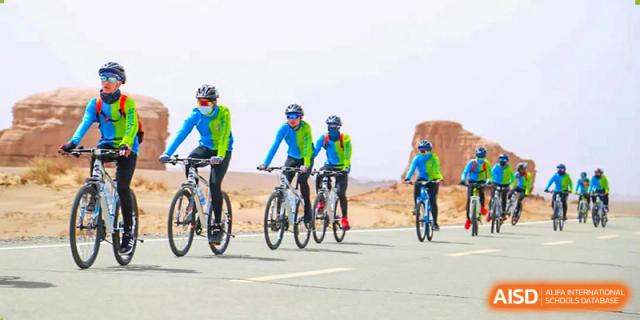 A group of students from Guangdong Country Garden School in blue and yellow gear ride bicycles on a road in a desert-like landscape.