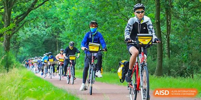 Students from Guangdong Country Garden School wearing helmets and masks ride bicycles on a path surrounded by trees.