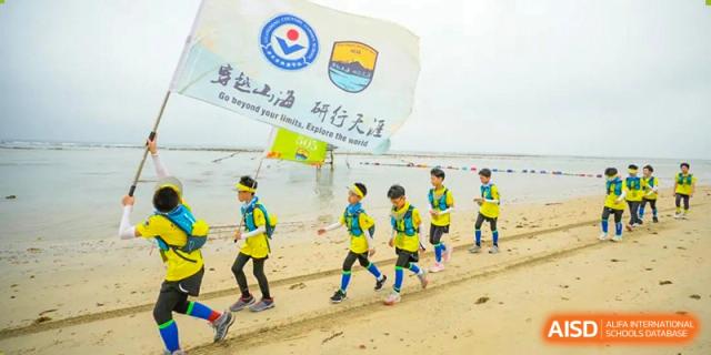 Students from Guangdong Country Garden School in blue and yellow uniforms hold a banner on a sandy beach.