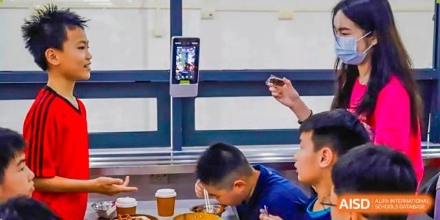 A young male student from Guangdong Country Garden School sits at a table while a man uses a thermometer for a health check.