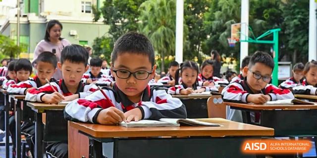 A young male student from Shenzhen Yaohua Experimental School wearing glasses, focused on writing at his desk in an outdoor learning setting.