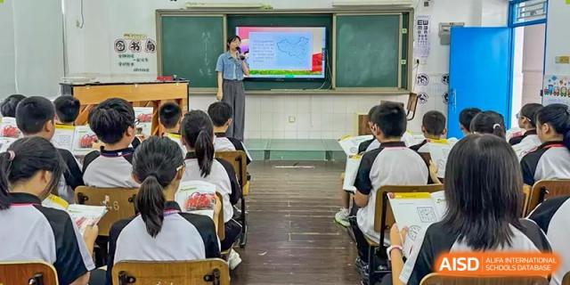 A female teacher at Shenzhen Yaohua Experimental School lecturing to students in a classroom with a large presentation screen.