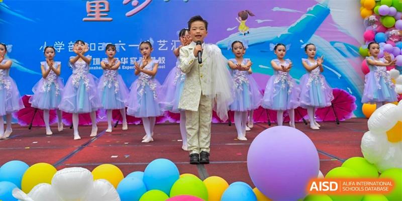 A young male student from Shenzhen Yaohua Experimental School singing into a microphone on stage, surrounded by students in white and blue costumes.