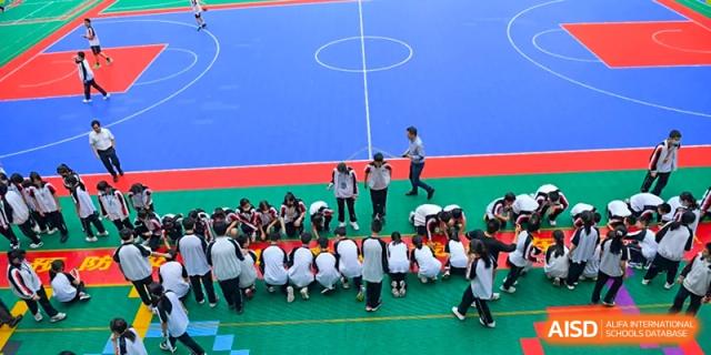 Shenzhen Yaohua Experimental School students seated on the ground in a huddle while teachers supervise an outdoor activity on a blue sports court.