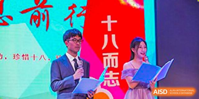 A teacher or student speaking on a microphone in front of a red banner at Huzhou South Taihu Bilingual School.