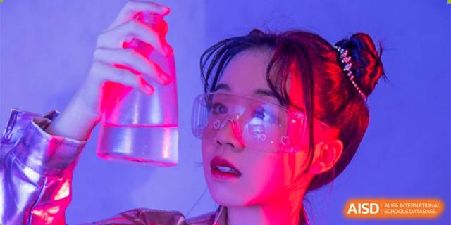 A female student from The First High School of Changsha, Hunan wearing safety goggles and holding a test tube under bright, colorful laboratory lighting.