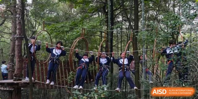 Students from The First High School of Changsha, Hunan navigating a high ropes course outdoors, wearing safety harnesses.
