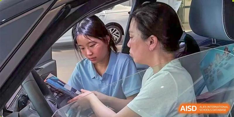 Two female students or a student and teacher from The First High School of Changsha, Hunan sitting together in a car or indoor setting, looking down at a mobile device.