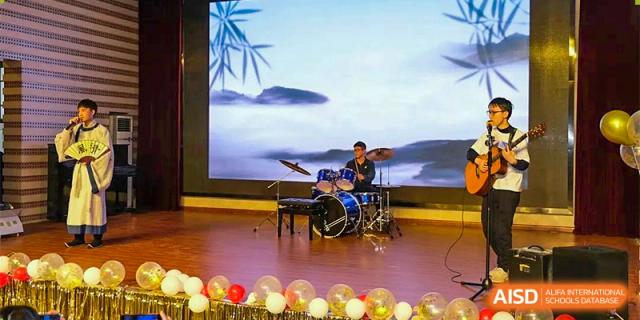 A student from The First High School of Changsha, Hunan playing a drum set and keyboard on a stage with a scenic landscape backdrop and balloons in front.