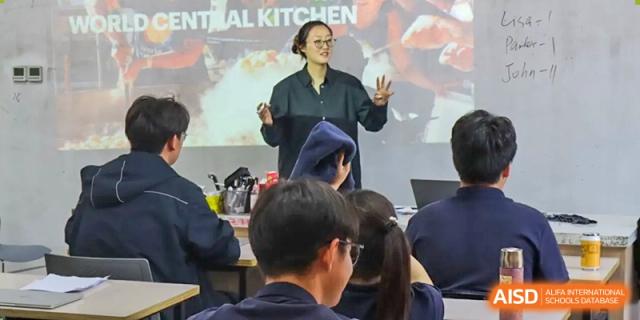 A Vindu-Charlie Lakefield College instructor or chef giving a demonstration in front of a class with a screen displaying "AI KITCHEN."