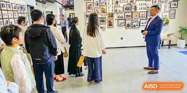 A female Vindu-Charlie Lakefield College student in a white jacket looks at a wall display of photos and artwork in a gallery or hallway.