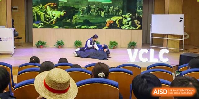 A performer on a stage at Vindu-Charlie Lakefield College kneeling or bowing in traditional attire, with an audience watching from their seats.