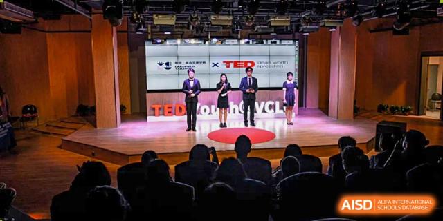 Three speakers from Vindu-Charlie Lakefield College standing on a red-lit stage giving a presentation to an audience in a darkened auditorium.