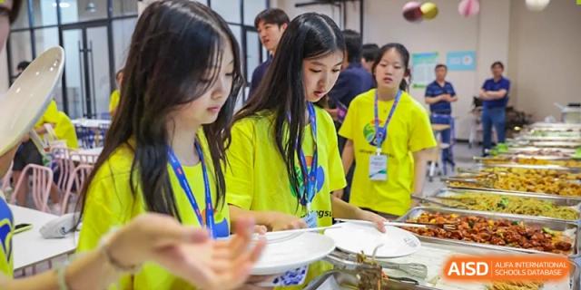 A female student in a yellow shirt serves herself food from a buffet line, demonstrating a meal service at Vindu-Charlie Lakefield College.