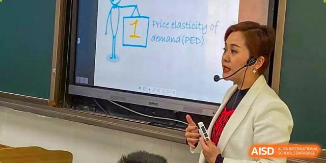 A teacher from The High School Attached to Hunan Normal University explaining "Price elasticity of demand (PED)" using a presentation on a smart board.