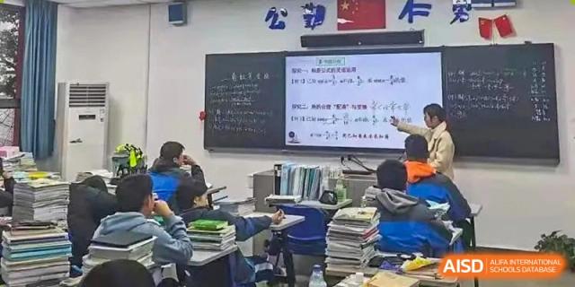A busy classroom at The High School Attached to Hunan Normal University with students working at their desks and complex equations written on a traditional blackboard.