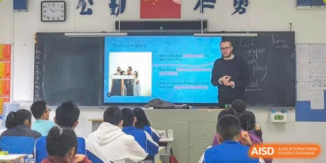 A teacher from The High School Attached to Hunan Normal University giving a lesson using a projected slide about "Shark Tank" to students seated at desks.