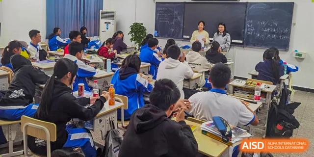 A teacher from The High School Attached to Hunan Normal University interacting with a wide class of students seated at traditional desks in a classroom setting.