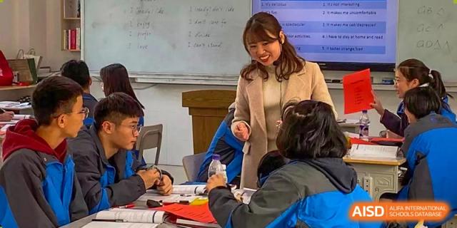 A female teacher from The High School Attached to Hunan Normal University engaging directly with a small group of students at their desks in the classroom.