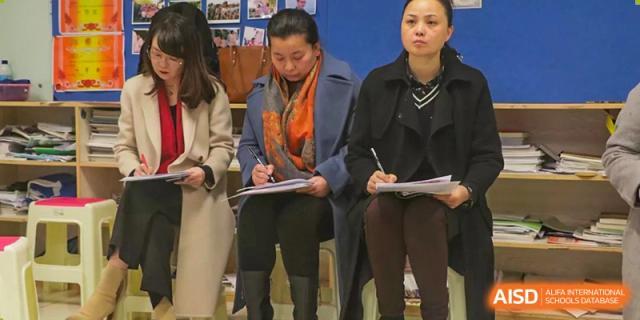 Several adults, potentially staff or observers from The High School Attached to Hunan Normal University, standing and taking notes on clipboards during a lesson or presentation.