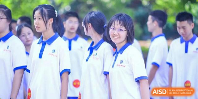 A smiling female student from The High School Attached to Hunan Normal University wearing a white and blue uniform, with other blurred students in the background.