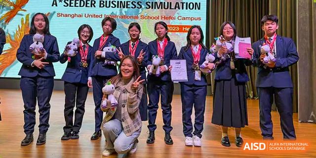 Group photo of students from The High School Attached to Hunan Normal University on stage, smiling and posing with awards and bouquets after a Business Simulation event.