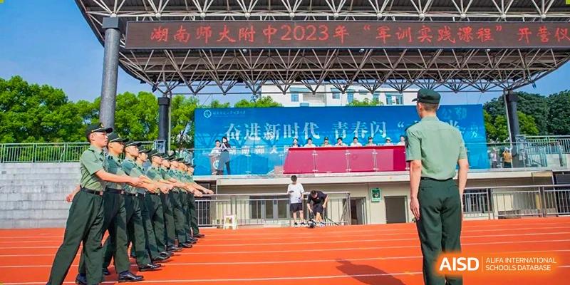 Students from The High School Attached to Hunan Normal University on a running track and field, with a large banner celebrating the 2023 "Military Training" event in the background.