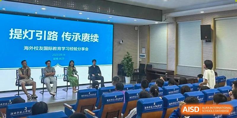 Students from The High School Attached to Hunan Normal University attending a lecture or seminar in a tiered auditorium, with two speakers seated on the stage.
