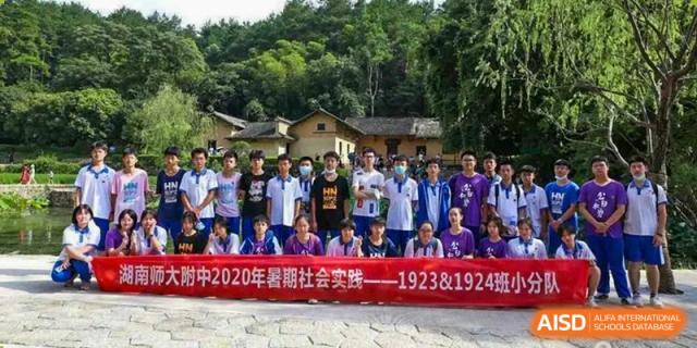 A large group of students from The High School Attached to Hunan Normal University posing outdoors in a rural setting with traditional buildings, possibly during a social practice trip.