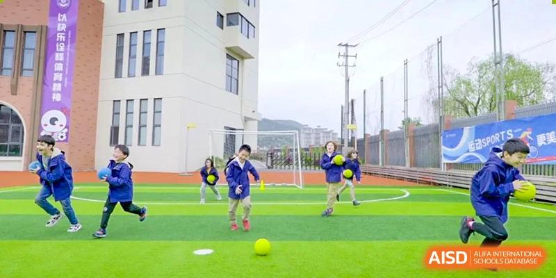 Young Wenzhou Union International School (WUIS) students in blue jackets playing with a yellow ball on a bright green outdoor field.