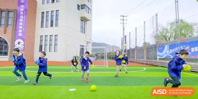Young Wenzhou Union International School (WUIS) students in blue jackets playing with a yellow ball on a bright green outdoor field.