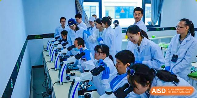 CNUH-GA Curriculum Partnership Program A group of students in white lab coats and safety glasses working side-by-side at a long bench with microscopes in a laboratory.