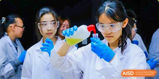 CNUH-GA Curriculum Partnership Program A close-up of a student in a lab coat and safety glasses, wearing blue gloves, carefully pouring a yellow liquid from one container into another.