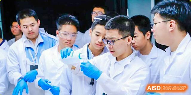 CNUH-GA Curriculum Partnership Program Three students in white lab coats, goggles, and blue gloves closely examining a clear liquid in a glass container.