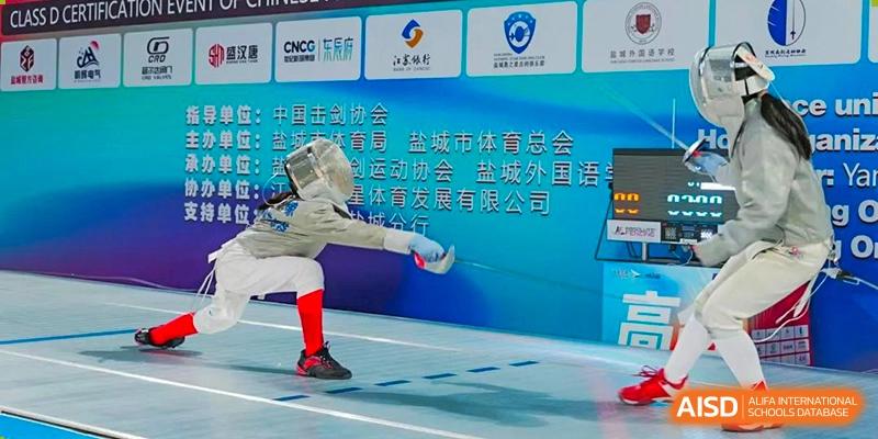 A close-up shot of a fencer in action during a competition, with a backdrop showing Chinese text for a sporting event sponsored by Yancheng Foreign Language School and other organizations.