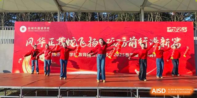 Two female Yancheng Foreign Language School performers in red outfits dancing on an outdoor stage with a large red banner backdrop.