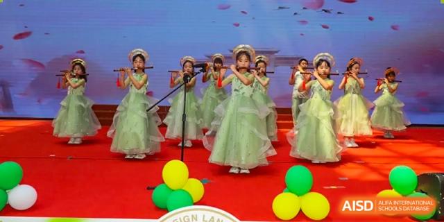 A group of Yancheng Foreign Language School students performing a song or dance on stage, all wearing flowing green dresses, with colorful balloons around them.