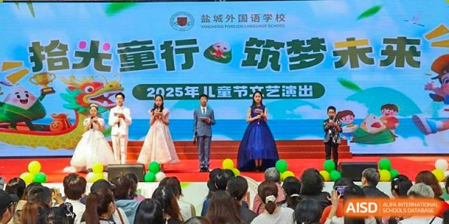 Two adults on a stage addressing an audience of Yancheng Foreign Language School students during a children's festival or performance event, with a 2025 banner visible.