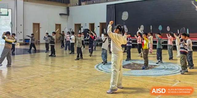 A Yancheng Foreign Language School student raising their hands in a gesture of success or effort while practicing a group physical exercise in an indoor hall.