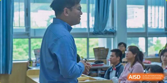 A male teacher or instructor facing a classroom, speaking during a lesson near a window, Fuyuan British American School.