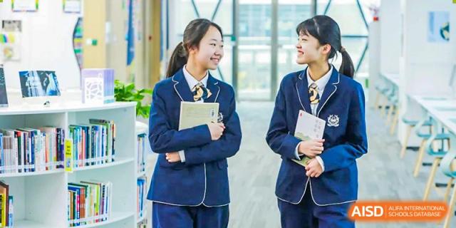 Two female students in dark blue school uniforms with yellow ties standing and smiling in a bright school hallway at ISA Wuhan International School.