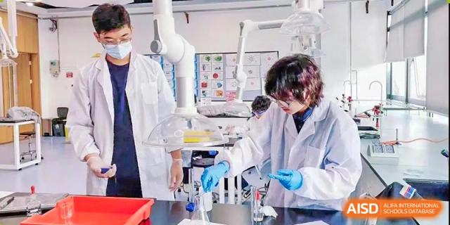 Two students in white lab coats and blue gloves performing an experiment using glassware and lab equipment at ISA Wuhan International School.