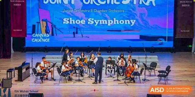 Students in an orchestra playing string instruments on a stage under a banner that reads "Junior Orchestra & Chamber Orchestra Shoe Symphony" at ISA Wuhan International School.