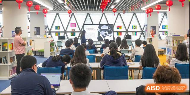 Students sitting in a brightly lit modern classroom with red lanterns hanging from the ceiling, looking at a screen displaying a historical figure at ISA Wuhan International School.