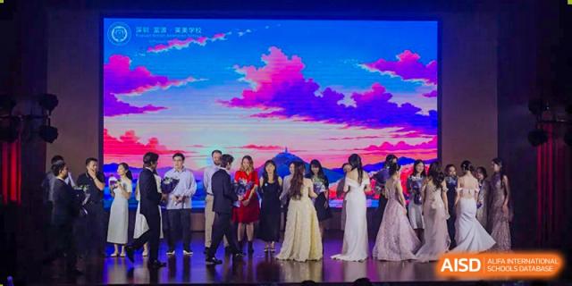 Students from Fuyuan British American School dressed in formal attire attending a gala or event on a stage with a brightly lit screen.