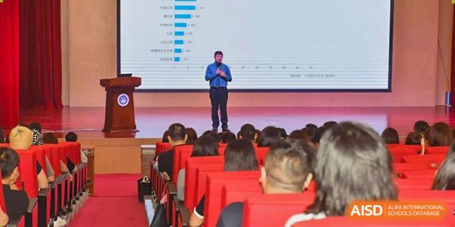 A speaker standing on a stage, addressing an audience of students and staff at a Fuyuan British American School assembly.